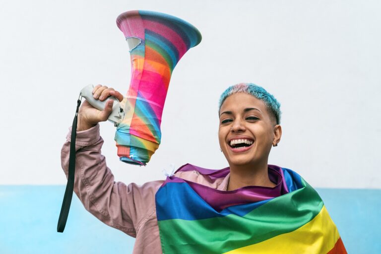 Person with a megaphone with rainbow colors on it and draped in a Pride flag