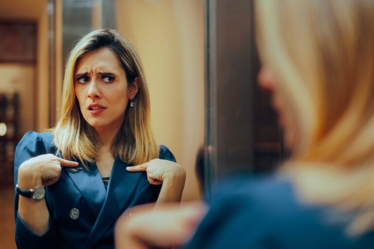 Woman looking in mirror and pointing at herself with questioning look on her face