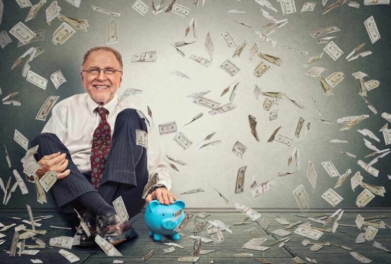 Older man sitting on the floor with blue piggie bank and dollars falling down around him