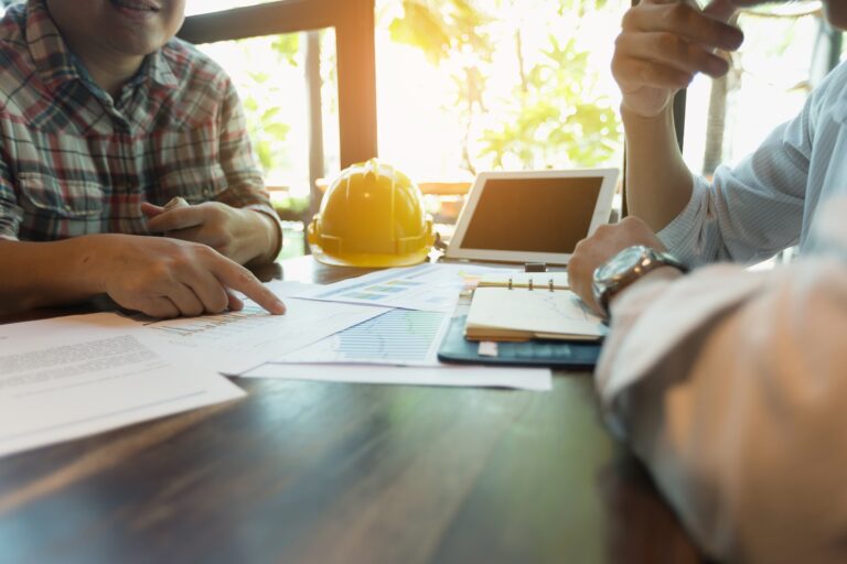 Two people sitting at a table with papers and a hard hat in front of them