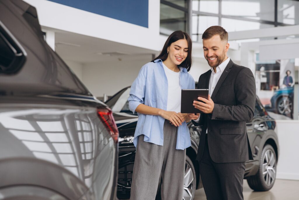 Man in a suite talking to woman dressed in light blue button up and slacks with purse on her shoulder, standing next to a car