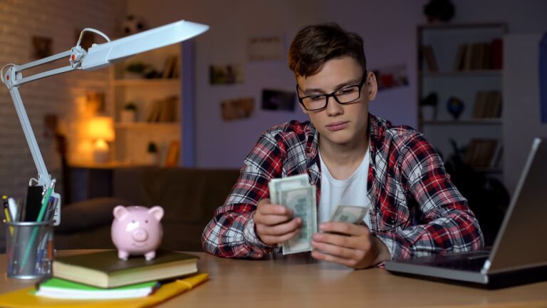 Young man in glasses, counting dollar bills at his desk. Desk has lamp and papers on it.